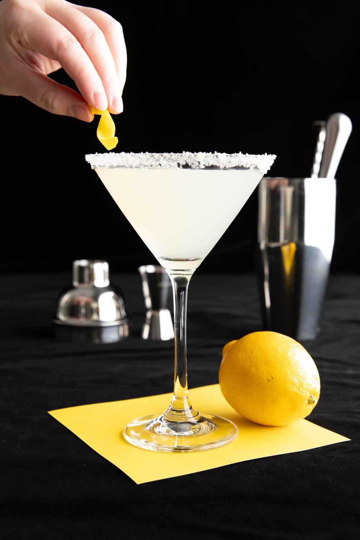 Hand platziert eine Zitronenzeste auf dem Zitronen-Zucker-Rand eines Lemon Drops.