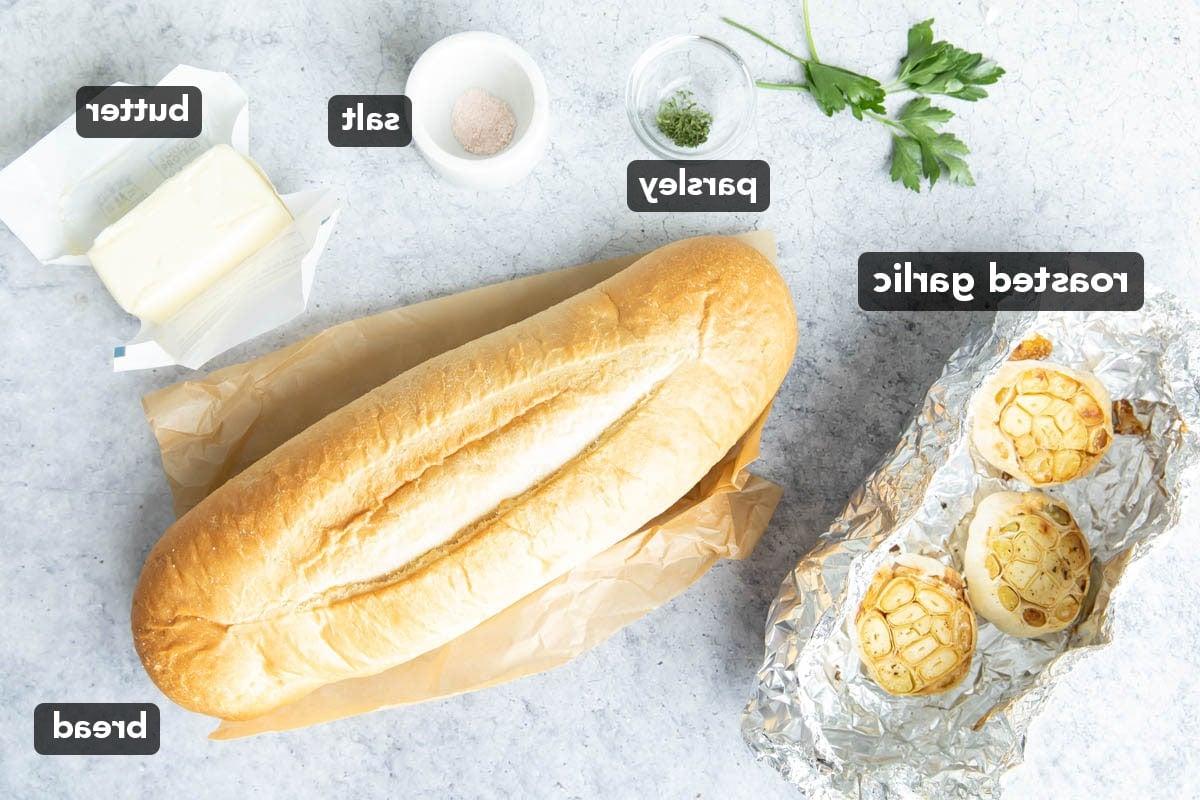 Zutaten für geröstetes Knoblauchbrot, darunter ein Brotlaib, Butter, Salz, gerösteter Knoblauch und Petersilie auf einem Küchentisch