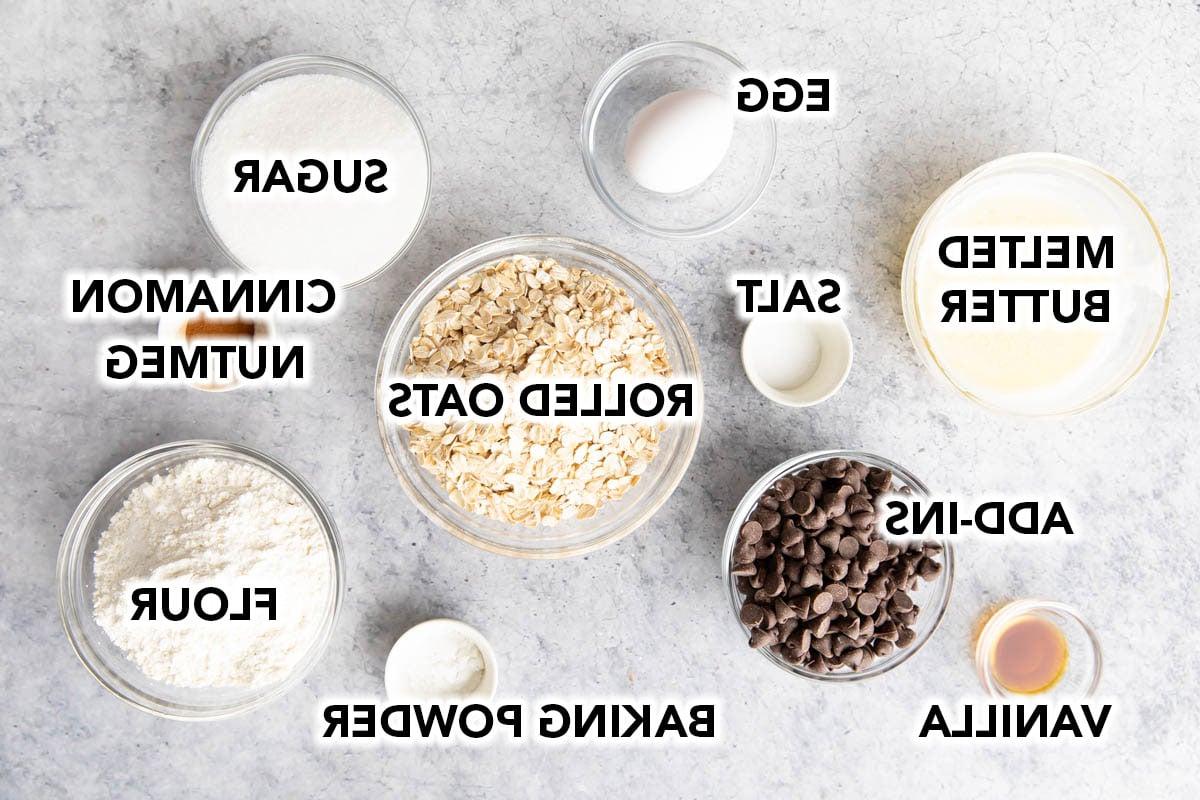 Die Zutaten für Haferflocken-Cookie-Riegel in vorbereiteten Schalen, darunter Haferflocken, Mehl, Zimt, Ei, Salz, Butter, Schokostückchen, Zucker, Vanilleextrakt und Backpulver.