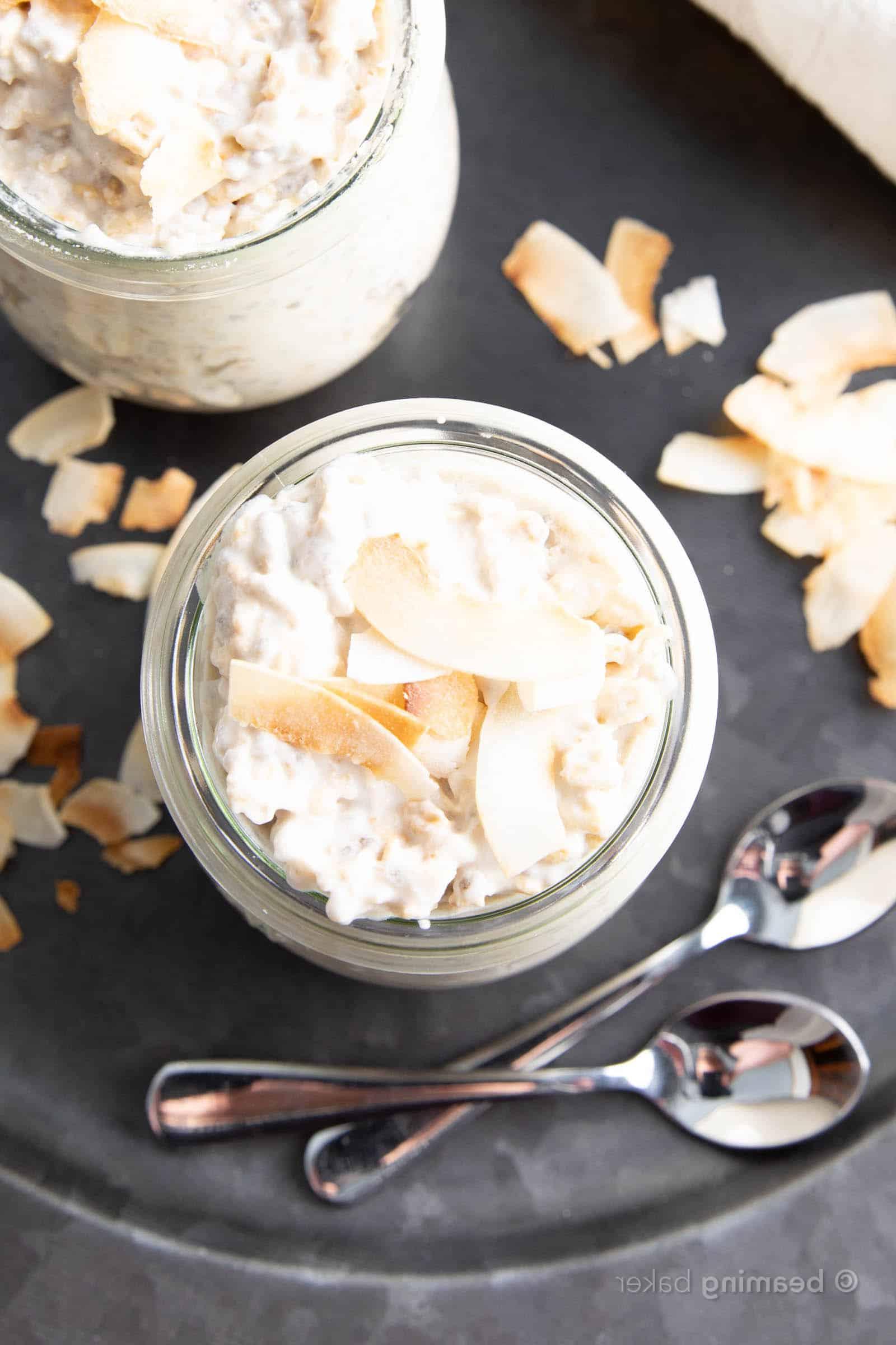 Nahaufnahme von oben von zwei Gläsern Kokos Overnight Oats mit zwei Löffeln und passendem Geschirr