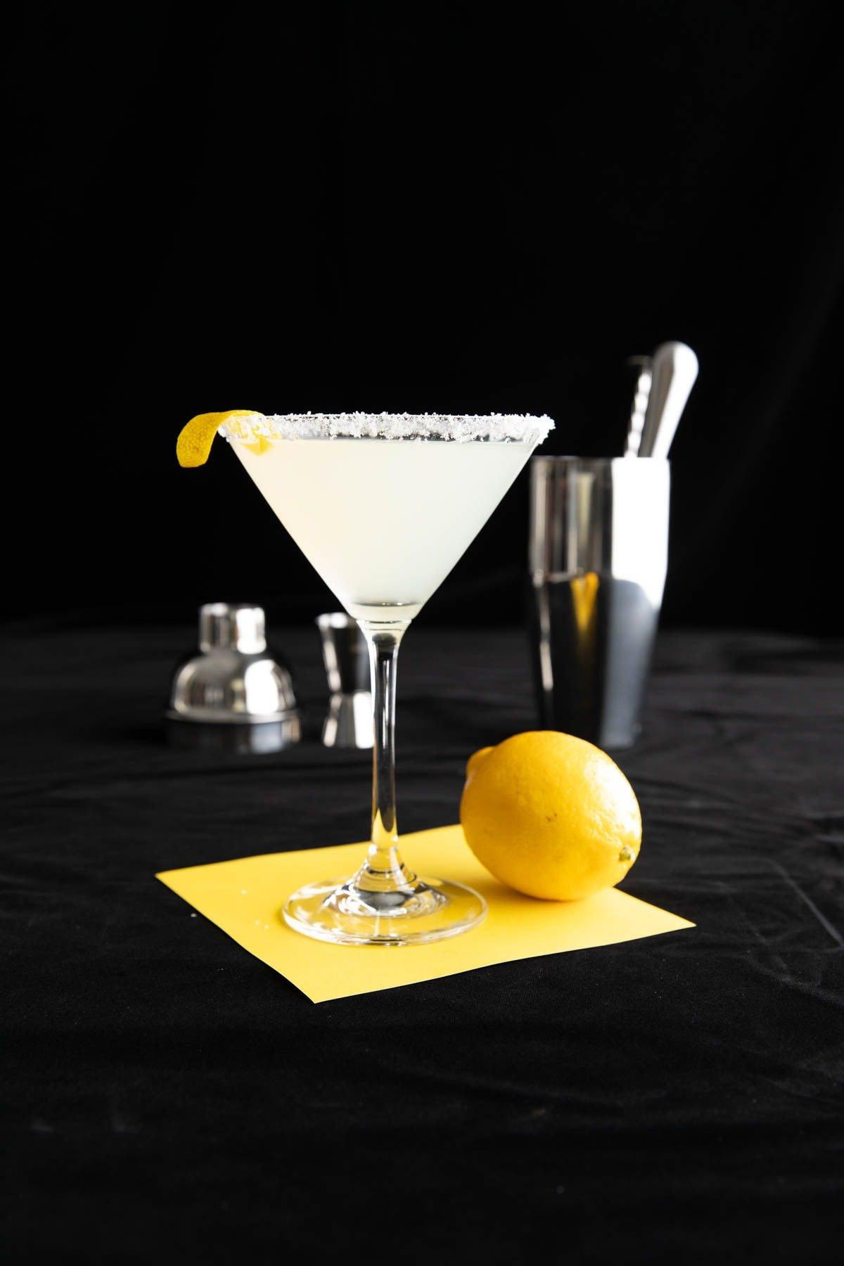 Dieses Cocktailrezept wird in einem Cocktailglas mit Zuckerrand und einem Cocktailset im Hintergrund serviert.