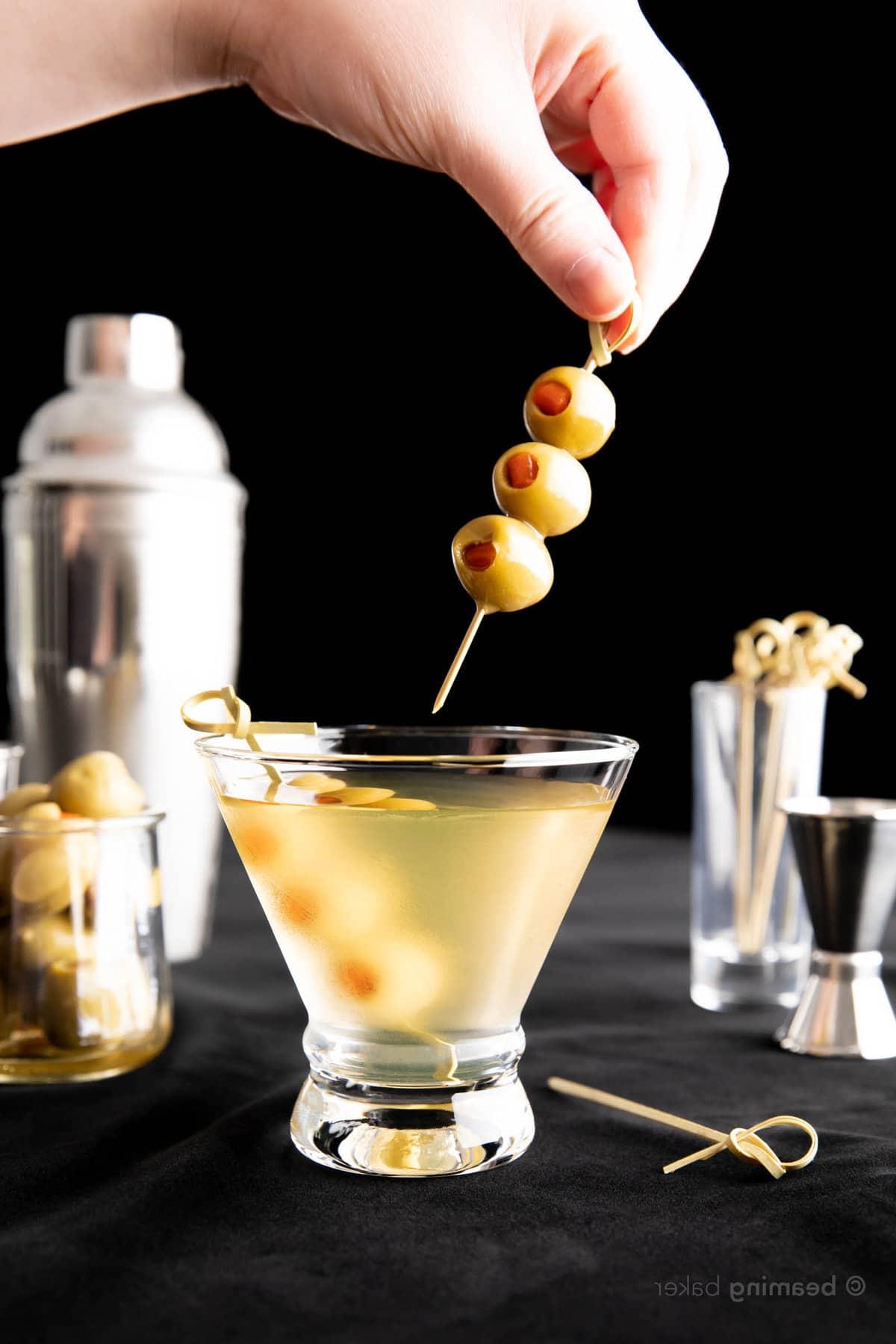 Extra Schmutziger Martini 8 Hand hält einen zweiten Oliven-Spieß, um den Extra Dirty Martini vor schwarzem Hintergrund zu vervollständigen